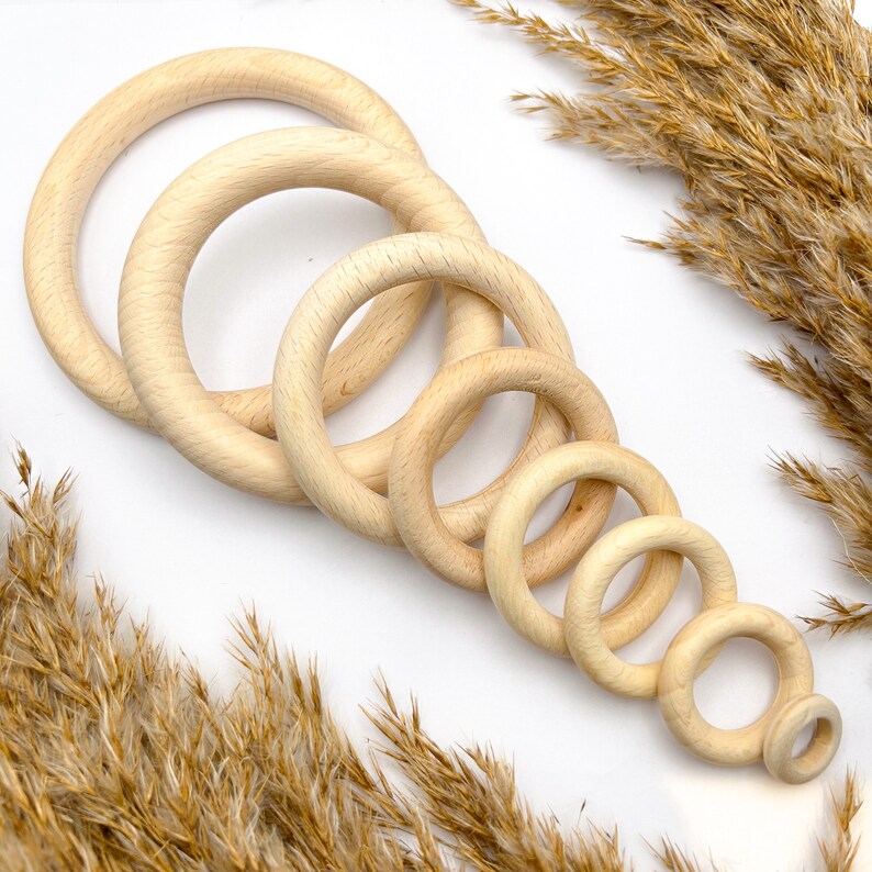 May include: A collection of six unfinished wooden rings of varying sizes, arranged diagonally. The rings are light brown and smooth, with a natural wood grain. The smallest ring has a small wooden peg attached. Dried wheat stalks are in the background.