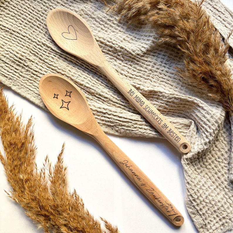 May include: Two wooden spoons with engraved sayings. One spoon has a heart and the text "Bei Mama schmeckt's am besten". The other spoon has stars and the text "Johanna & Zacharias".