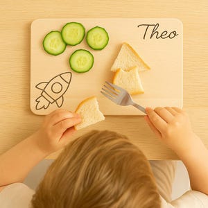 Peut inclure: Planche &agrave; d&eacute;couper en bois avec le nom "Theo" en &eacute;criture noire, orn&eacute;e d'une illustration de fus&eacute;e. Des tranches de concombre et du pain sont dispos&eacute;s sur la planche. Un enfant tient un morceau de pain et une fourchette.