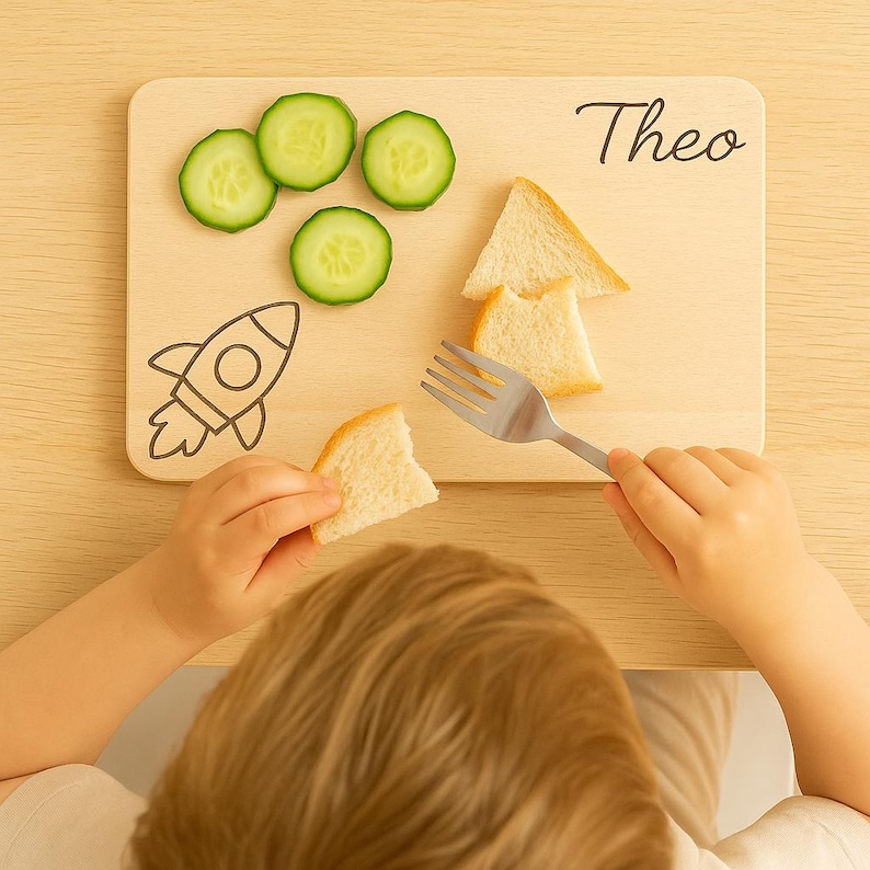 Peut inclure: Planche &agrave; d&eacute;couper en bois avec le nom "Theo" en &eacute;criture noire, orn&eacute;e d'une illustration de fus&eacute;e. Des tranches de concombre et du pain sont dispos&eacute;s sur la planche. Un enfant tient un morceau de pain et une fourchette.