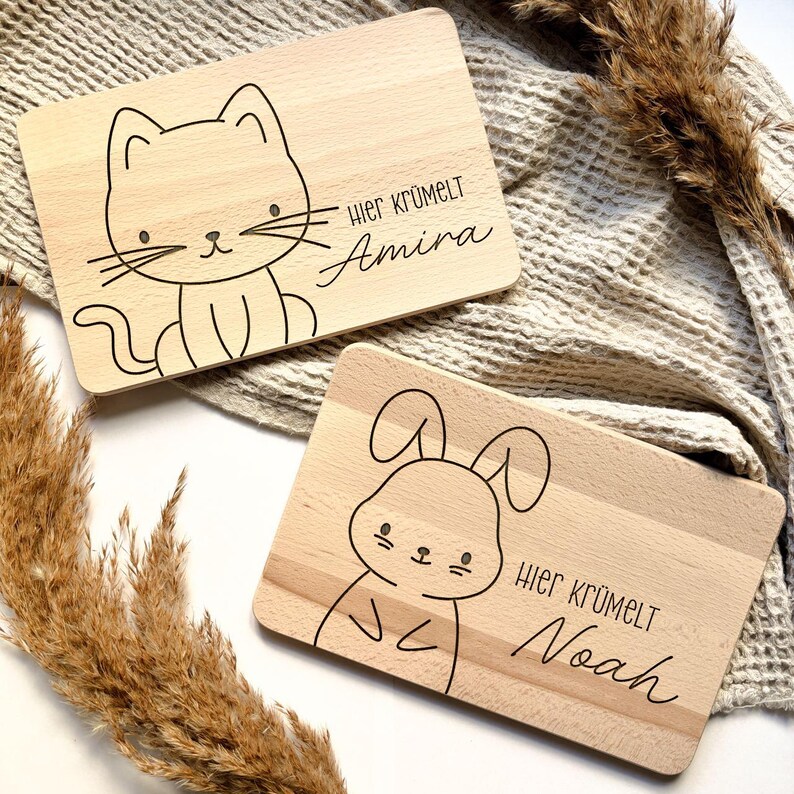 May include: Two wooden cutting boards with engraved animal illustrations and text. The first board features a cat with the text "Hier kr&uuml;melt Amira". The second board features a rabbit with the text "Hier kr&uuml;melt Noah".