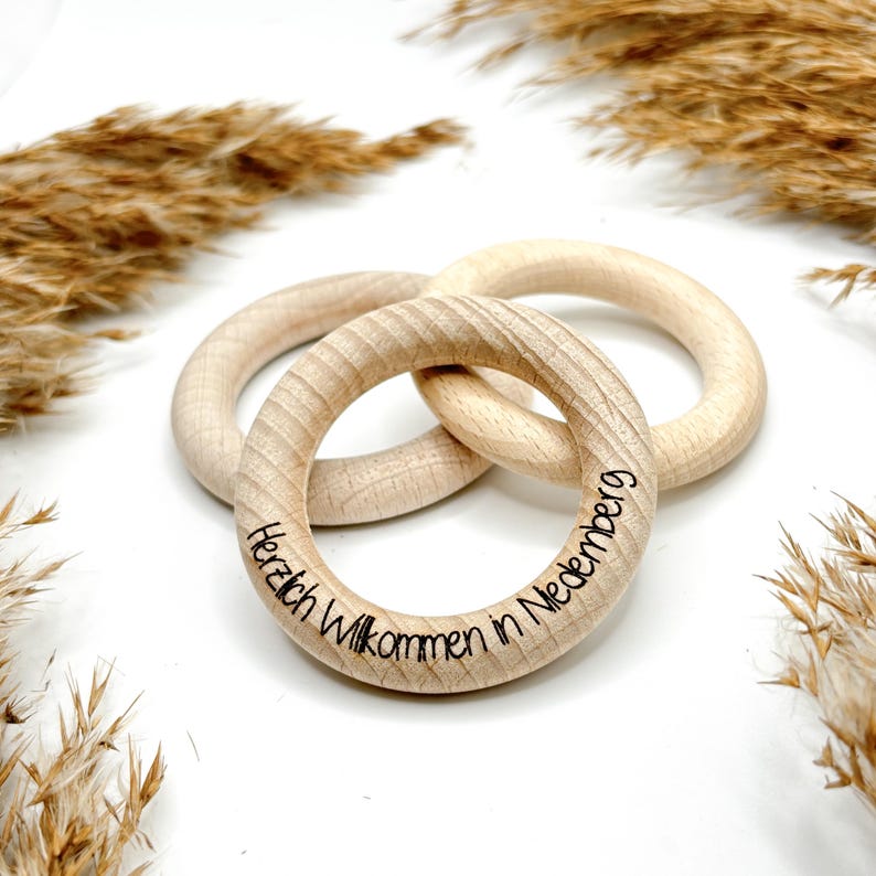 May include: Three wooden teething rings interlocked, made of light-colored wood. One ring has the engraved text "Herzlich Willkommen in Nedenberg". The rings are arranged on a white surface, with dried plants in the background.