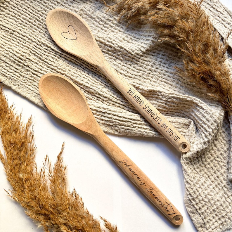 May include: Two wooden spoons with engraved text. One spoon says "Bei Mama schmeckt's am besten" and the other says "Johanna + Zach 2022". The spoons are on a beige fabric background.