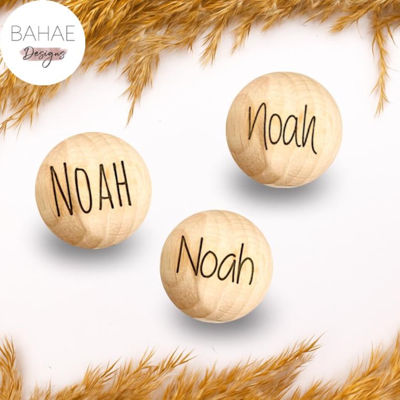 May include: Three wooden spheres with the name "Noah" printed in black. The spheres are light brown and sit on a white surface, with dried wheat stalks in the background. The top left corner has a logo that says "BAHAE Designs".