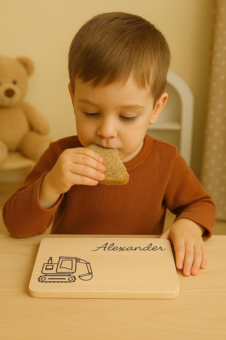 Peut inclure: Un enfant en pull marron mange du pain devant une planche en bois. La planche porte le nom "Alexander" et un dessin de pelle m&eacute;canique. Un ours en peluche est en arri&egrave;re-plan.