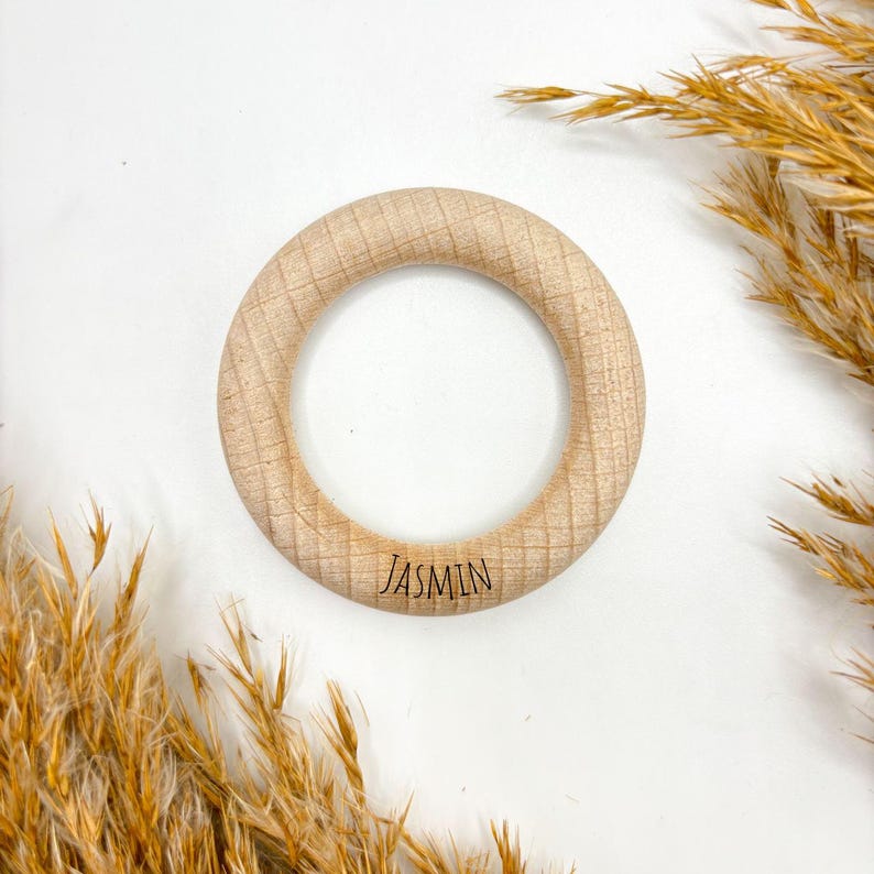 Könnte beinhalten: Ein hellbrauner Beißring aus Holz mit dem eingravierten Namen "JASMIN". Der Ring hat eine strukturierte Oberfläche. Der Hintergrund ist weiß mit getrockneten Pflanzen.