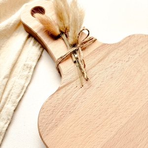May include: A light-colored wooden cutting board with a handle and a decorative bundle of dried flowers tied with twine. The board has a unique, curved shape and a smooth surface. The flowers are beige and brown.