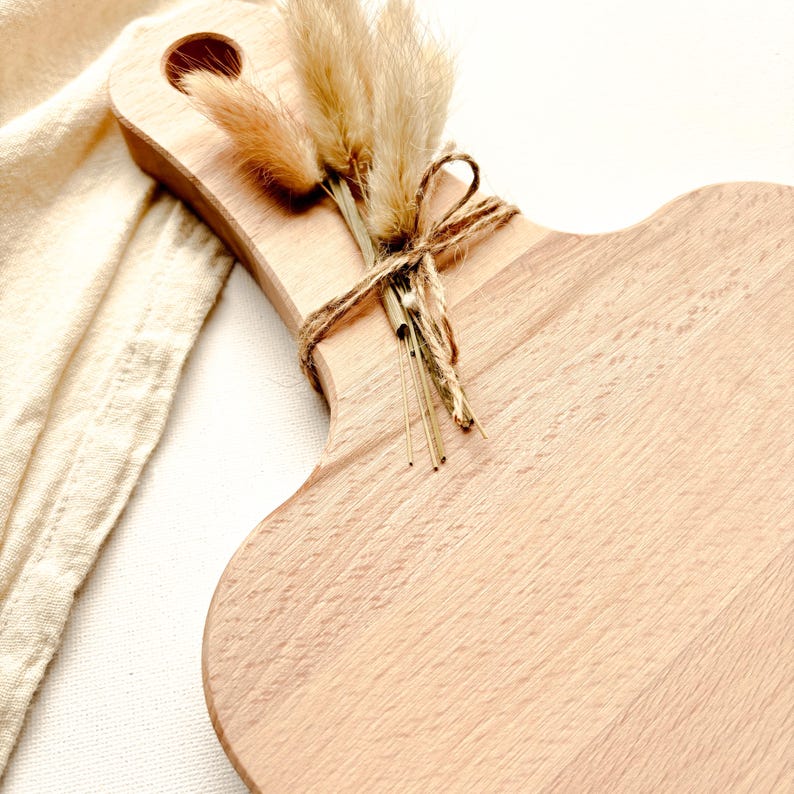 May include: A light-colored wooden cutting board with a handle and a decorative bundle of dried flowers tied with twine. The board has a unique, curved shape and a smooth surface. The flowers are beige and brown.