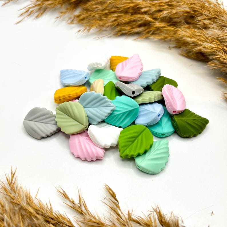 Könnte beinhalten: Eine Sammlung von silikonförmigen Blätterperlen in verschiedenen Pastellfarben, darunter Grün, Blau, Rosa und Grau. Die Perlen haben ein Loch in der Mitte zum Aufziehen.