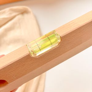 May include: A wooden level with a clear, bubble-filled vial. The level is made of light-colored wood and has a yellow and green liquid inside the vial. The level is designed to help ensure surfaces are straight and even.