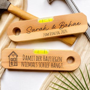 May include: Two wooden levels with yellow vials, personalized with text. One reads "Sarah & Bahae ZUM EINZUG 2025", the other has a house graphic and text. Hammers and fabric are in the background.