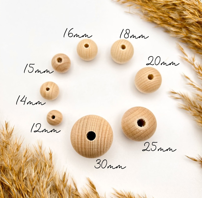 May include: A collection of unfinished wooden beads in various sizes, ranging from 12mm to 30mm in diameter. The beads are arranged on a white surface, with measurements in millimeters written next to each bead. Dried grasses frame the composition.