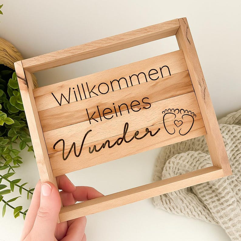 May include: A light-colored wooden sign with the words "Willkommen kleines Wunder" in black script. The sign features a rectangular frame and a design of baby footprints with a heart. The sign is held by a person's hand.