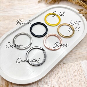 May include: A close-up of six different key rings in various finishes: silver, gunmetal, black, gold, light gold, and rose gold. The key rings are arranged in a circle on a white surface.