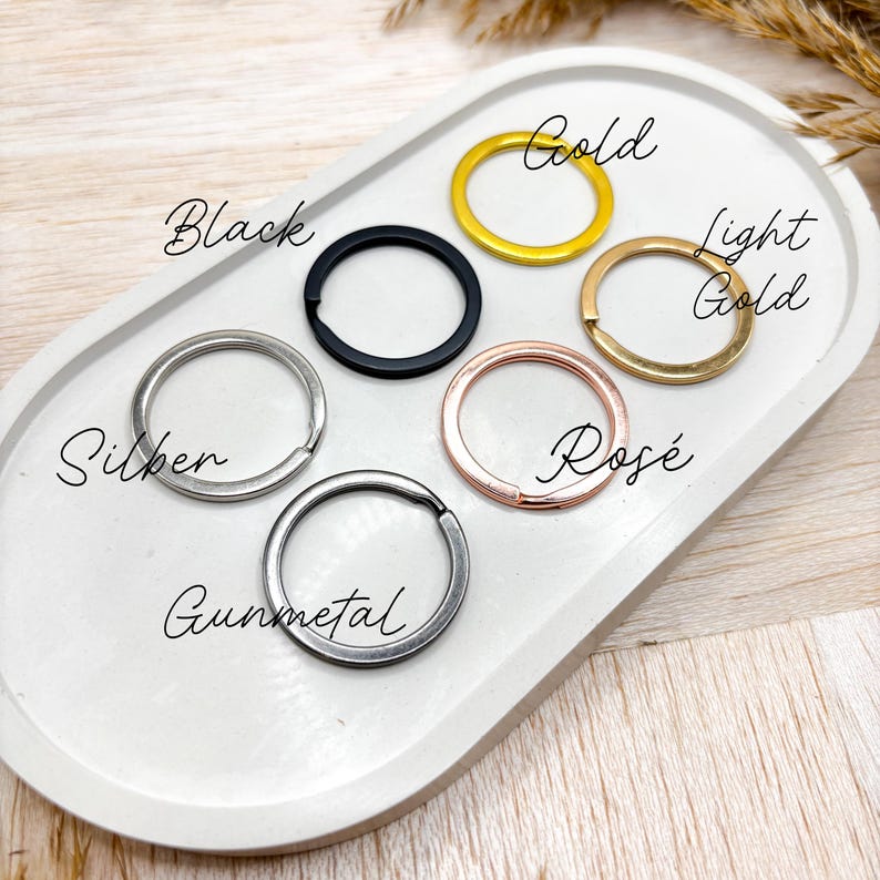 May include: A close-up of six different key rings in various finishes: silver, gunmetal, black, gold, light gold, and rose gold. The key rings are arranged in a circle on a white surface.