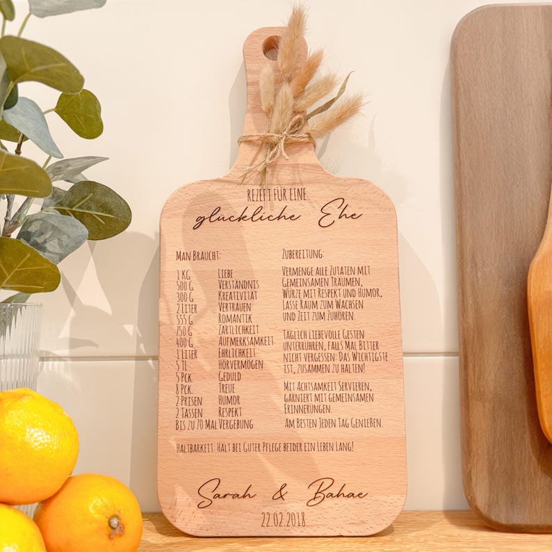 Könnte beinhalten: Holzschneidebrett mit Gravur eines Rezepts für eine glückliche Ehe. Es zeigt eine Zutatenliste und Anweisungen, die Namen "Sarah & Bahac" und das Datum "22.02.2018". Dekoriert mit Trockenblumen.