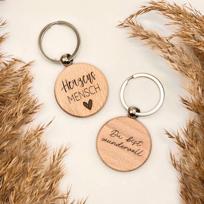 May include: Two wooden keychains with silver key rings. One keychain reads "Herzensmensch" with a heart symbol. The other keychain reads "Du bist wundervoll". The keychains are round and made of light-colored wood.