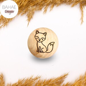 Può includere: Pomello rotondo in legno con un disegno di volpe inciso al laser. La volpe è raffigurata con linee nere in stile cartone animato. Il pomello è marrone chiaro. L'immagine include anche il testo "BAHAE Designs" in un cerchio bianco.