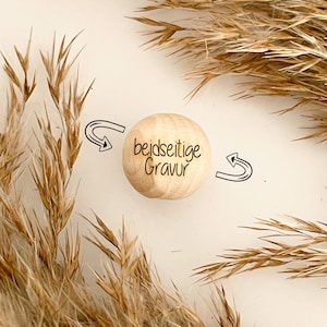 May include: A light-colored wooden sphere with the text "beidseitige Gravur" in black script. The sphere is surrounded by two black arrows and dried plants. The background is a neutral off-white color.