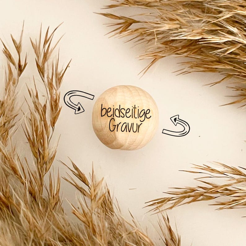 May include: A light-colored wooden sphere with the text "beidseitige Gravur" in black script. The sphere is surrounded by two black arrows and dried plants. The background is a neutral off-white color.