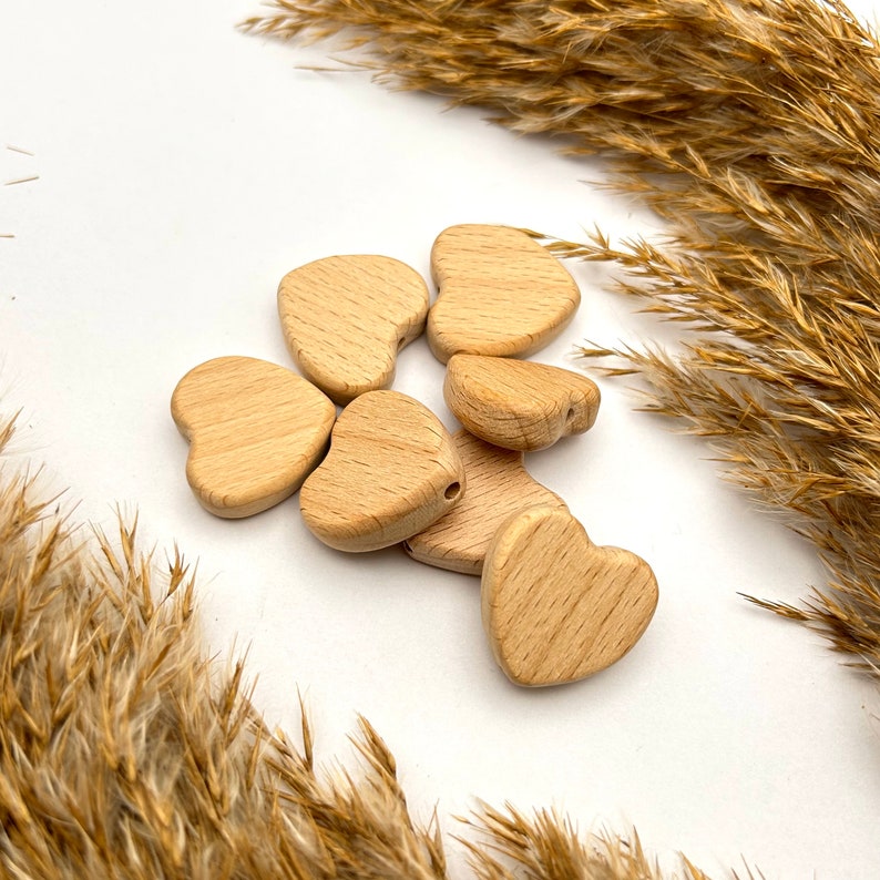 May include: A close-up of six natural wooden heart-shaped beads. The beads are arranged in a pile on a white surface. The beads are smooth and have a light brown color.