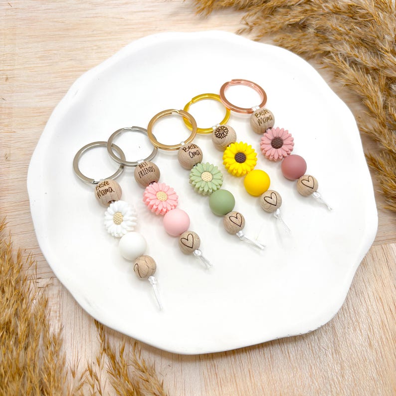 Könnte beinhalten: Fünf Schlüsselanhänger mit Holzperlen, Herzanhängern und Blumenanhängern. Die Anhänger sind in Pink, Grün, Gelb und Weiß. Die Schlüsselanhänger sind mit "Best Mom", "Best Friend", "Best Oma", "Best Nana" und "Best Grandma" beschriftet.