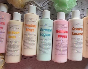 May include: Seven cylindrical bottles of body wash from The Breezy Beachwood Co. with white caps, featuring labels titled Whipped Dream, Sun Your Buns, Tropical Blast, Mermaid Lagoon, Rainbow Crush, Creamy Coconut, and Citrus Flame, with bath sponges.