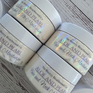 Könnte beinhalten: Mehrere Tiegel Gesichts- und Halsfeuchtigkeitscreme liegen auf einer Holzoberfläche. Die Tiegel haben weiße Deckel und schillernde Etiketten mit dem Text "ALOE AND PEARL Face and Neck Moisturizer" und "The Breezy Beachwood Co."