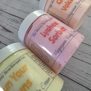 K&ouml;nnte beinhalten: Drei zylindrische Tiegel mit Magnesium-K&ouml;rpercreme und wei&szlig;en Deckeln. Die Etiketten tragen die Aufschrift The Breezy Beachwood Co und zeigen die Duftrichtungen Citrus Volcano in Orange, Lychee Sorbet in Rosa und Sun Your Buns in Gelb.