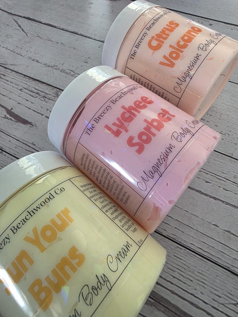K&ouml;nnte beinhalten: Drei zylindrische Tiegel mit Magnesium-K&ouml;rpercreme und wei&szlig;en Deckeln. Die Etiketten tragen die Aufschrift The Breezy Beachwood Co und zeigen die Duftrichtungen Citrus Volcano in Orange, Lychee Sorbet in Rosa und Sun Your Buns in Gelb.