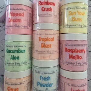 K&ouml;nnte beinhalten: Acht zylindrische Kunststoffdosen mit Magnesium-K&ouml;rpercreme von The Breezy Beachwood Co. mit wei&szlig;en Deckeln. Die Etiketten zeigen verschiedene Duftnamen wie Rainbow Crush, Whipped Dream, Sun Your Buns, Tropical Blast, Cucumber Aloe, Raspberry Mojito, Citrus Volcano, Fresh Powder und Lychee Sorbet, jeweils mit einem Nettogewicht von 227g auf der Seite.