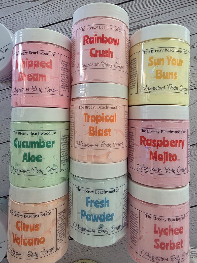 K&ouml;nnte beinhalten: Acht zylindrische Kunststoffdosen mit Magnesium-K&ouml;rpercreme von The Breezy Beachwood Co. mit wei&szlig;en Deckeln. Die Etiketten zeigen verschiedene Duftnamen wie Rainbow Crush, Whipped Dream, Sun Your Buns, Tropical Blast, Cucumber Aloe, Raspberry Mojito, Citrus Volcano, Fresh Powder und Lychee Sorbet, jeweils mit einem Nettogewicht von 227g auf der Seite.