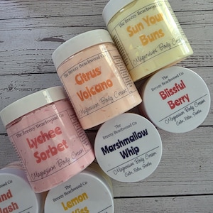 K&ouml;nnte beinhalten: Eine Sammlung runder Kunststoffdosen mit Magnesium-K&ouml;rpercreme von The Breezy Beachwood Co. Die Etiketten zeigen verschiedene Duftrichtungen wie Lychee Sorbet, Citrus Volcano, Sun Your Buns, Blissful Berry, Marshmallow Whip und Lemon Kiss.