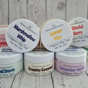 K&ouml;nnte beinhalten: Eine Sammlung runder Kunststoffdosen mit Magnesium-K&ouml;rpercreme von The Breezy Beachwood Co. Die Etiketten zeigen verschiedene Duftnamen, darunter Marshmallow Whip, Lemon Kiss, Blissful Berry, Coastal Storm, Creamy Coconut und Lavender Vanilla.