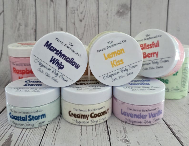 K&ouml;nnte beinhalten: Eine Sammlung runder Kunststoffdosen mit Magnesium-K&ouml;rpercreme von The Breezy Beachwood Co. Die Etiketten zeigen verschiedene Duftnamen, darunter Marshmallow Whip, Lemon Kiss, Blissful Berry, Coastal Storm, Creamy Coconut und Lavender Vanilla.
