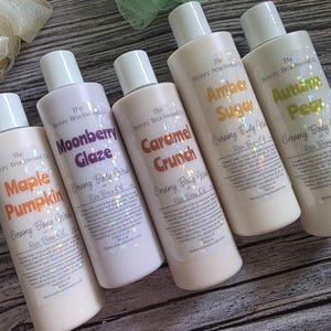 Könnte beinhalten: Fünf zylindrische Flaschen mit cremiger Körperwaschlotion von The Breezy Beachwood Co. mit Reiskleieöl. Die Etiketten zeigen holografischen Text für die Düfte Maple Pumpkin, Moonberry Glaze, Caramel Crunch, Amber Sugar und Autumn Pear.