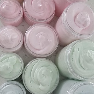 K&ouml;nnte beinhalten: Eine Sammlung von durchsichtigen Kunststoffdosen, gef&uuml;llt mit aufgeschlagener K&ouml;rperbutter in verschiedenen Pastellt&ouml;nen wie Hellrosa, sanftem Pfirsich, Minzgr&uuml;n und Hellblau. Die dicke, cremige Substanz ist in jedem Beh&auml;lter mit sichtbaren Spitzen.