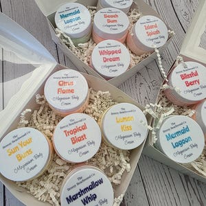 May include: Several small jars of Magnesium Body Cream are arranged in white boxes. Each jar has a white lid with a label featuring a unique name like "Marshmallow Whip" or "Mermaid Lagoon." The jars are nestled in white crinkle paper.
