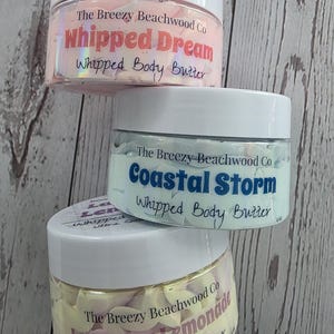 Könnte beinhalten: Drei durchsichtige Kunststoffdosen mit weißen Deckeln, die geschlagene Körperbutter enthalten. Die obere Dose ist rosa und mit Whipped Dream beschriftet, die mittlere ist hellblau mit Coastal Storm und die untere ist gelb und rosa mit Lavender Lemonade.