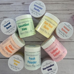 K&ouml;nnte beinhalten: Eine Sammlung von neun runden Kunststoffdosen mit Magnesium-K&ouml;rpercreme von The Breezy Beachwood Co. in verschiedenen Pastellfarben. Die Etiketten zeigen Duftnamen wie Lavender Vanilla, Coastal Storm, Cucumber Aloe, Sun Your Buns, Island Splash, Citrus Volcano, Lemon Kiss, Fresh Powder, Lychee Sorbet und Marshmallow Whip.