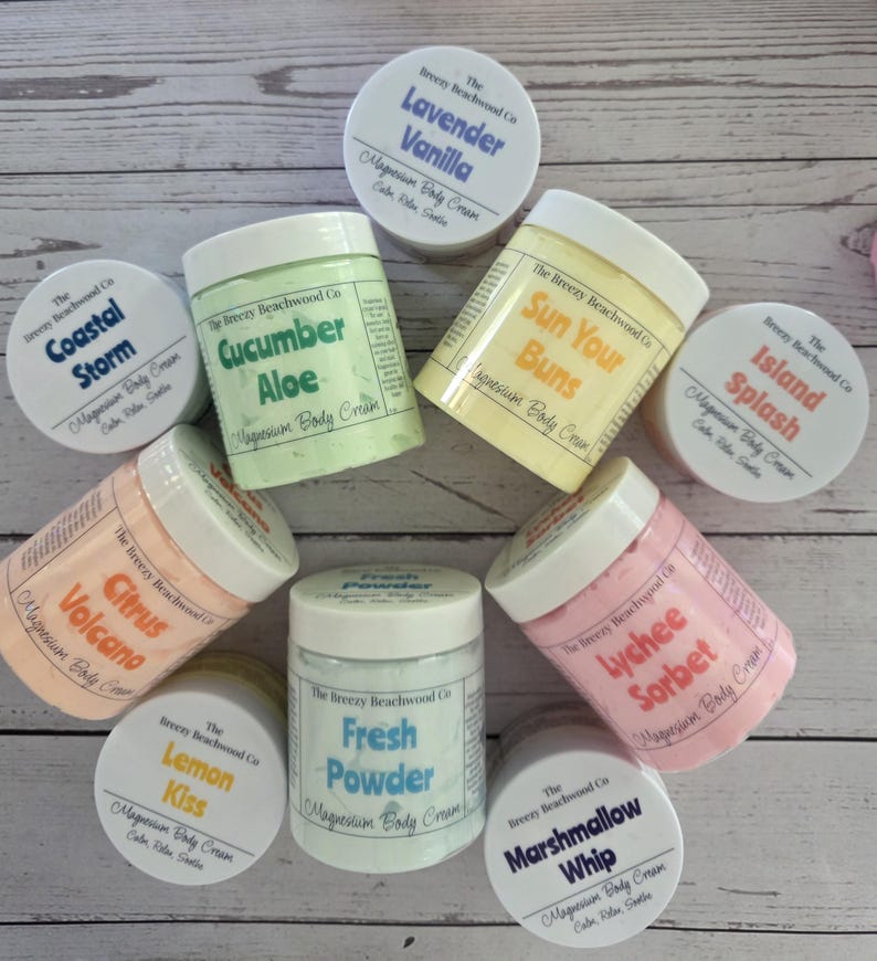 K&ouml;nnte beinhalten: Eine Sammlung von neun runden Kunststoffdosen mit Magnesium-K&ouml;rpercreme von The Breezy Beachwood Co. in verschiedenen Pastellfarben. Die Etiketten zeigen Duftnamen wie Lavender Vanilla, Coastal Storm, Cucumber Aloe, Sun Your Buns, Island Splash, Citrus Volcano, Lemon Kiss, Fresh Powder, Lychee Sorbet und Marshmallow Whip.