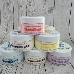 K&ouml;nnte beinhalten: Eine Sammlung von sechs runden Kunststoffdosen mit Magnesium-K&ouml;rpercreme von The Breezy Beachwood Co. in verschiedenen Pastellfarben. Die Etiketten tragen die Duftnamen Blissful Berry, Marshmallow Whip, Lemon Kiss, Coastal Storm, Creamy Coconut und Lavender Vanilla.