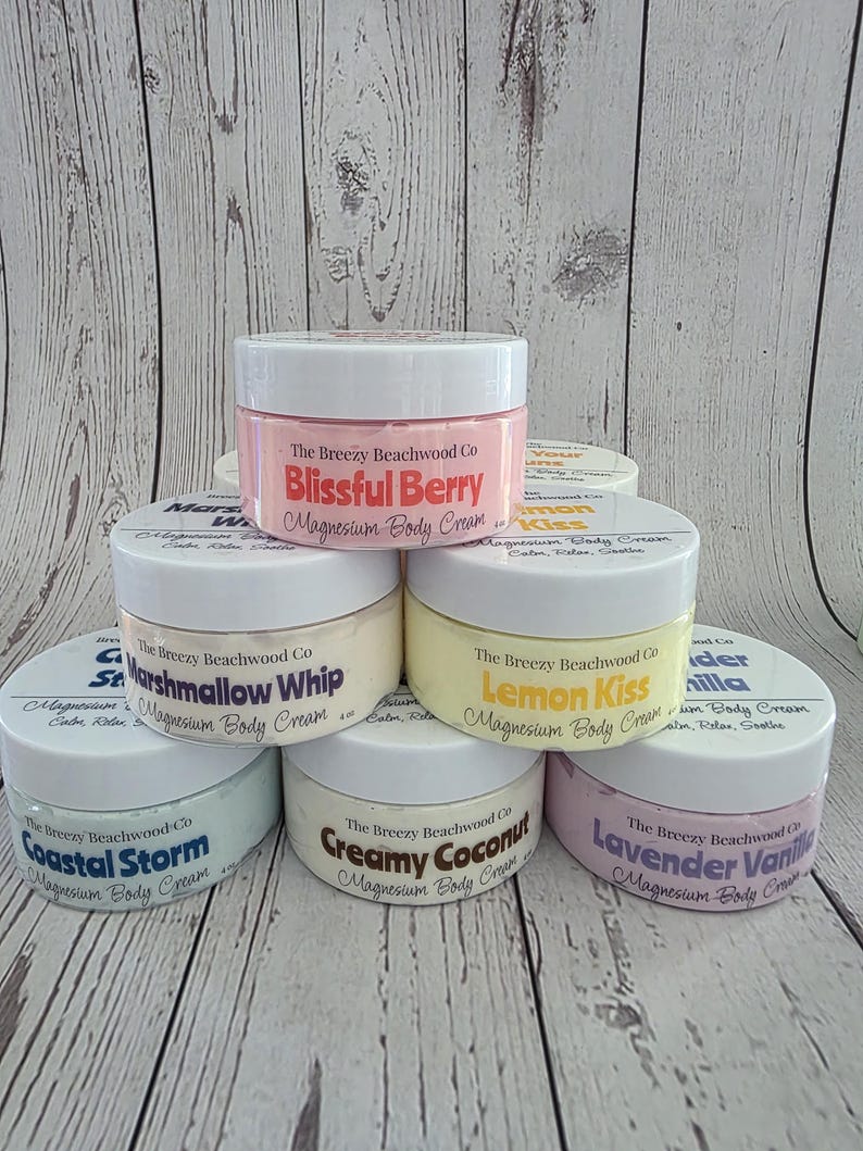 K&ouml;nnte beinhalten: Eine Sammlung von sechs runden Kunststoffdosen mit Magnesium-K&ouml;rpercreme von The Breezy Beachwood Co. in verschiedenen Pastellfarben. Die Etiketten tragen die Duftnamen Blissful Berry, Marshmallow Whip, Lemon Kiss, Coastal Storm, Creamy Coconut und Lavender Vanilla.
