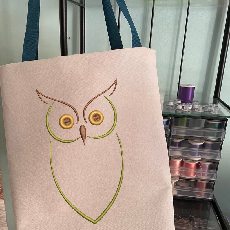 May include: A white canvas tote bag with a green and brown embroidered owl design. The owl has large, round eyes.