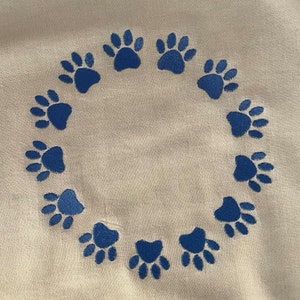 Puede incluir: Un fondo de tela blanco con un círculo de huellas de patas azules bordadas alrededor del centro.
