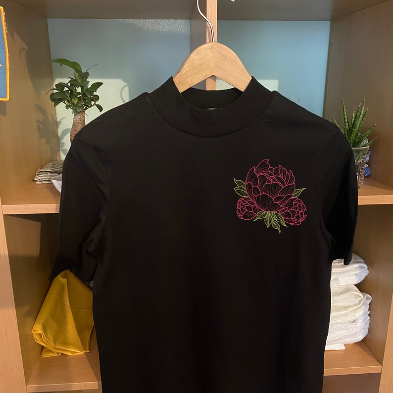 Pu&ograve; includere: Una maglietta nera a collo alto con un disegno floreale rosa e verde sul davanti. Il disegno floreale presenta un fiore di peonia con foglie verdi. La maglietta &egrave; appesa a una gruccia di legno.