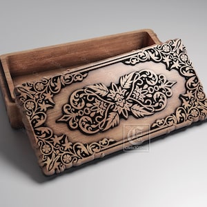 Puede incluir: Una caja de madera con tapa abierta, con intrincados tallados florales y de volutas. La caja es de madera marrón claro, con detalles marrón oscuro en el diseño tallado. El logotipo CNC es visible.