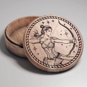 May include: A wooden box with a round lid. The lid is engraved with a detailed illustration of a woman with a bow and arrow, surrounded by stars. The box is open and the inside is visible.