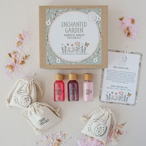 May include: A set of four small pouches containing pink and red bath salts, labelled "Fairy Crystals", "Morning Sunrise", "Dream Blast", and "Pixie Dust". The pouches are arranged on a white surface with a box labelled "Enchanted Garden Mindful Magic Potion Kit" and a card with the text "Once Upon a Time... Fairies and Pixies are powerful / loved / creative / joyful" in the background.