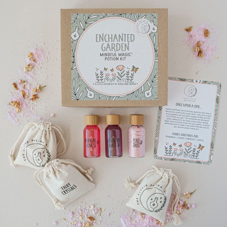 Puede incluir: Un conjunto de cuatro peque&ntilde;as bolsas que contienen sales de ba&ntilde;o rosas y rojas, etiquetadas como "Fairy Crystals", "Morning Sunrise", "Dream Blast" y "Pixie Dust". Las bolsas est&aacute;n dispuestas sobre una superficie blanca con una caja etiquetada como "Enchanted Garden Mindful Magic Potion Kit" y una tarjeta con el texto "&Eacute;rase una vez... Las hadas y los duendes son poderosos / amados / creativos / alegres" en el fondo.